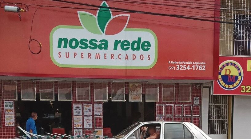 A fachada de um supermercado de bairro chamado 'Nossa Rede Supermercados'. Em destaque, um grande letreiro vermelho com o logotipo verde. Ao lado, uma placa da 'Drogaria Michele' e, em frente, a parte superior de um carro prateado.