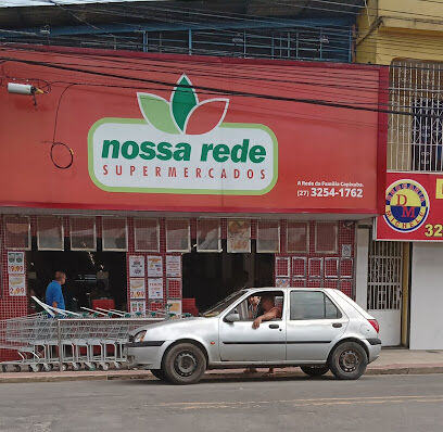 Fachada de um supermercado de bairro chamado 'Nossa Rede Supermercados'. Um carro prateado está estacionado na frente, e é possível ver cestos de compras na entrada. Ao lado, há uma placa com o nome de outra loja, 'Drogaria Michele'.