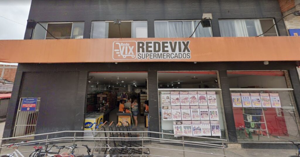 Fachada do Supermercado Rede Vix Jabaeté em Vila Velha. A imagem mostra a entrada principal com a placa "Rede Vix Supermercados", as vitrines com anúncios de ofertas e um carrinho de compras na frente da loja.